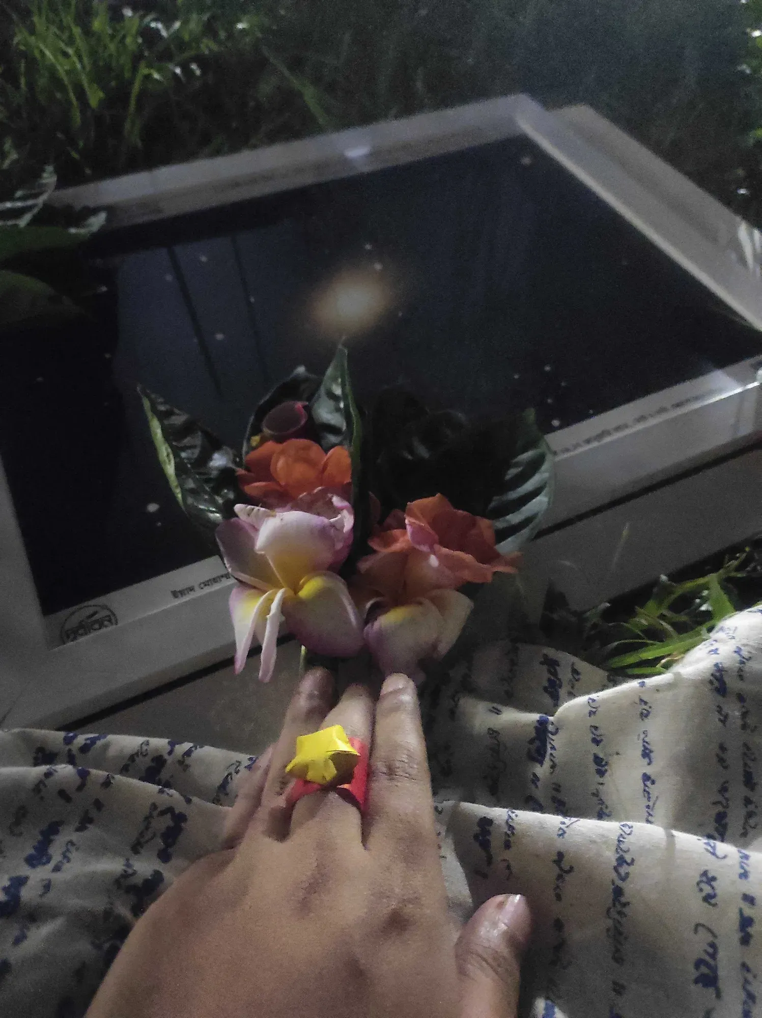 A hand wearing a handmade paper star ring, holding a small posy of flowers above a framed astrophotograph resting on the grass.