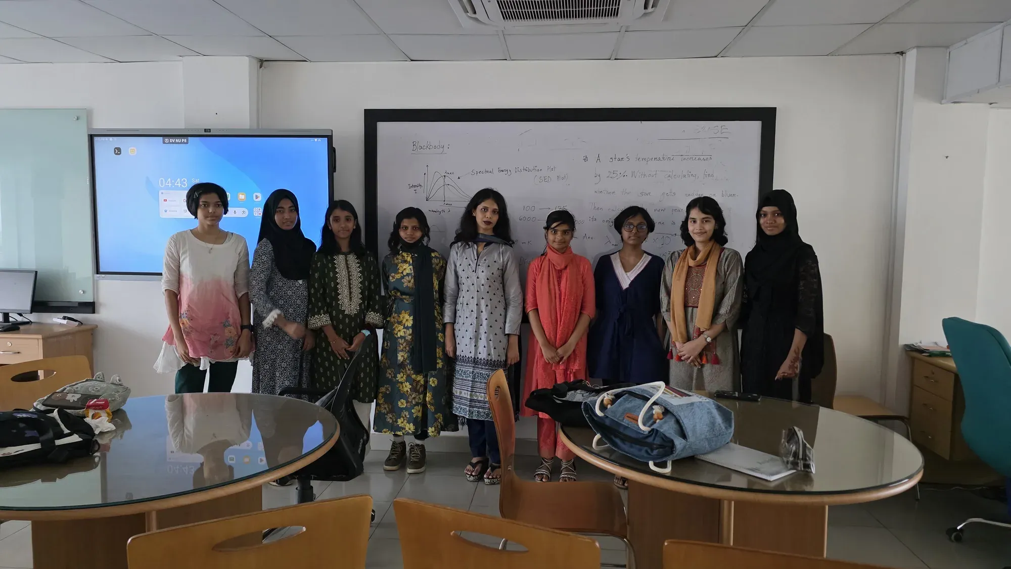 Participants of the BDOAA Women's Astronomy Olympiad Preparation Camp standing with an instructor in front of a whiteboard covered in blackbody and spectral energy distribution notes.