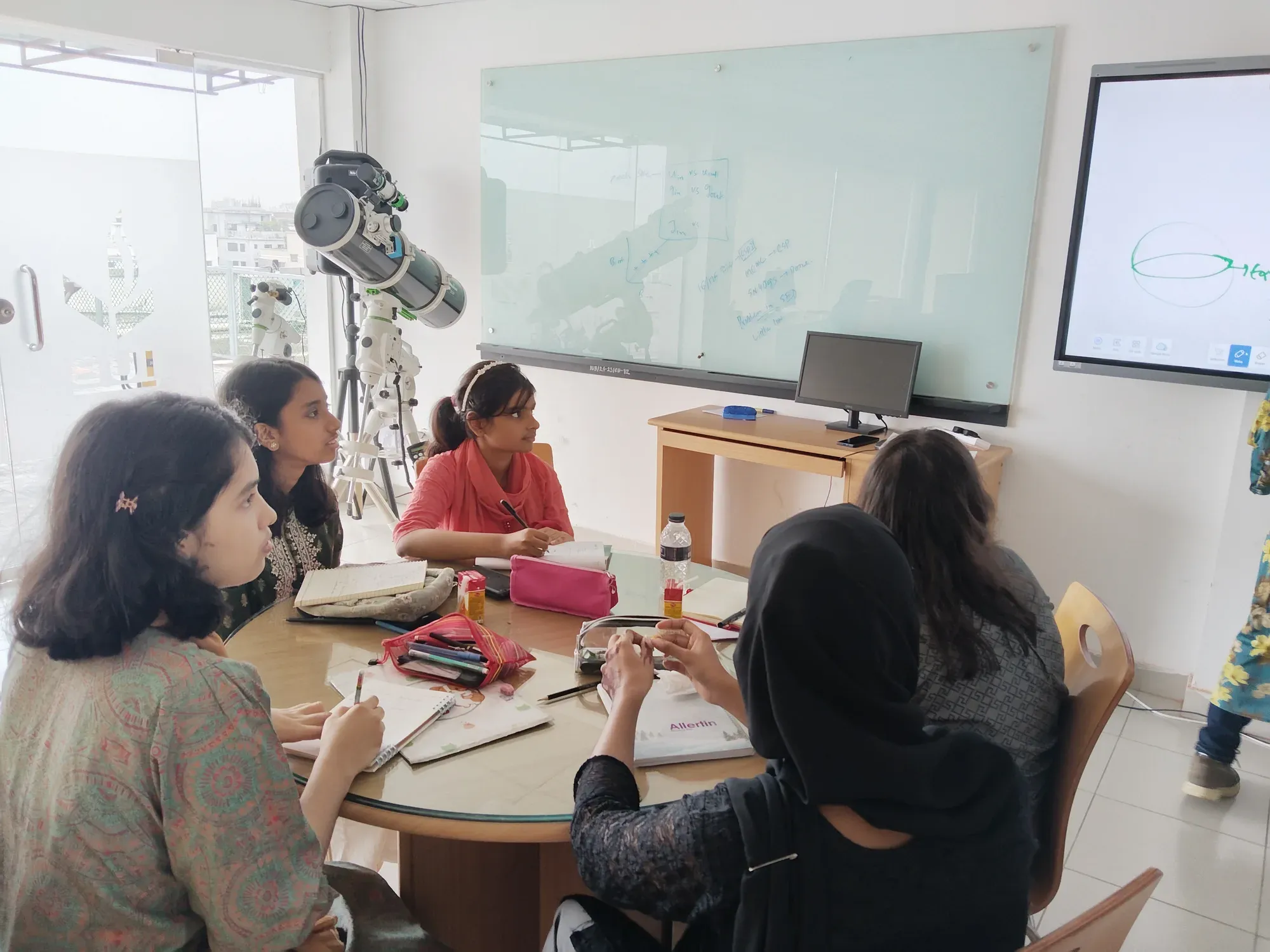Participants seated around a table looking towards the screen, with a large reflecting telescope set up beside the window.
