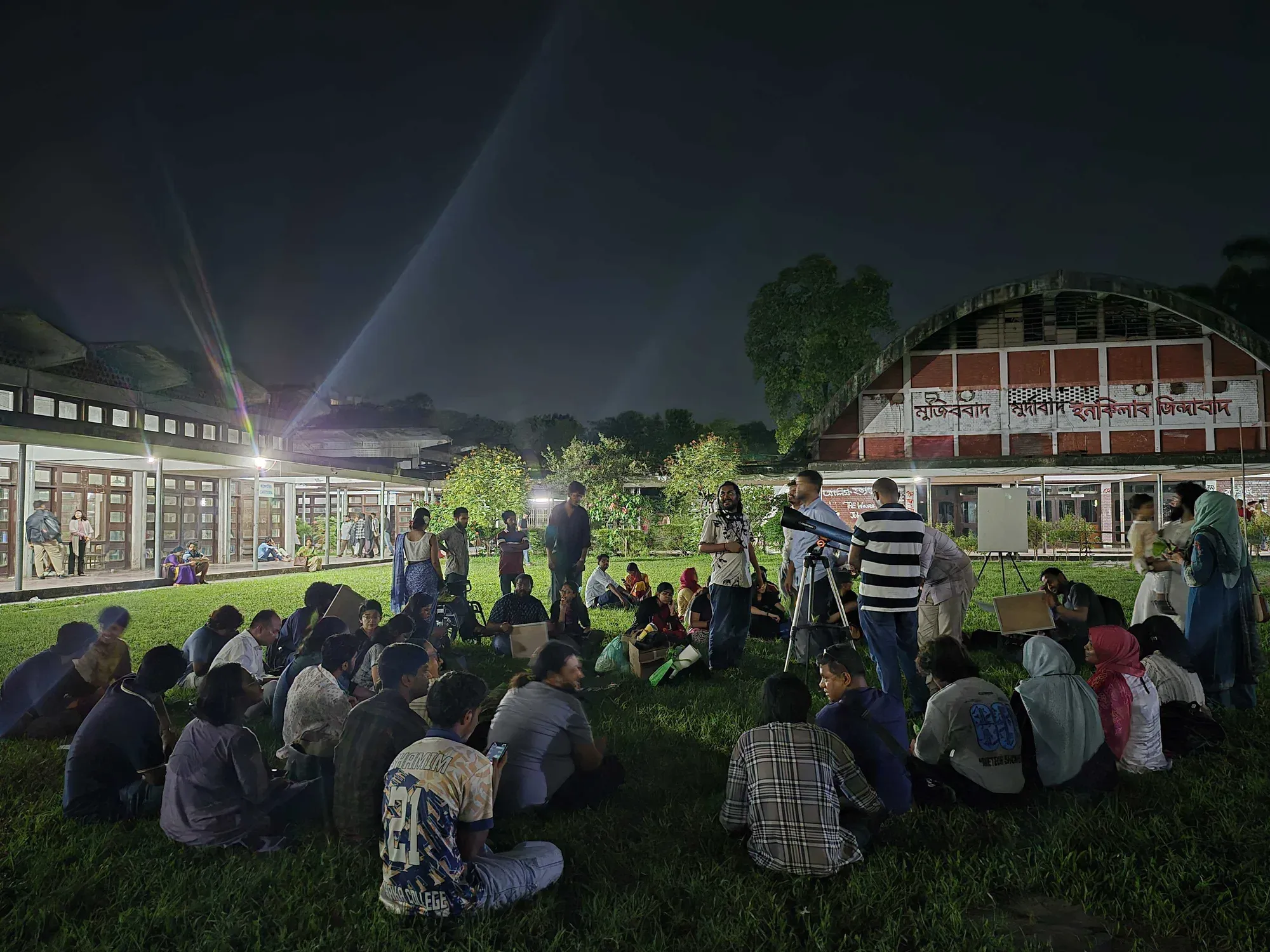 A large circle of participants seated on the grass at night, with a telescope and volunteers at the centre of the gathering.