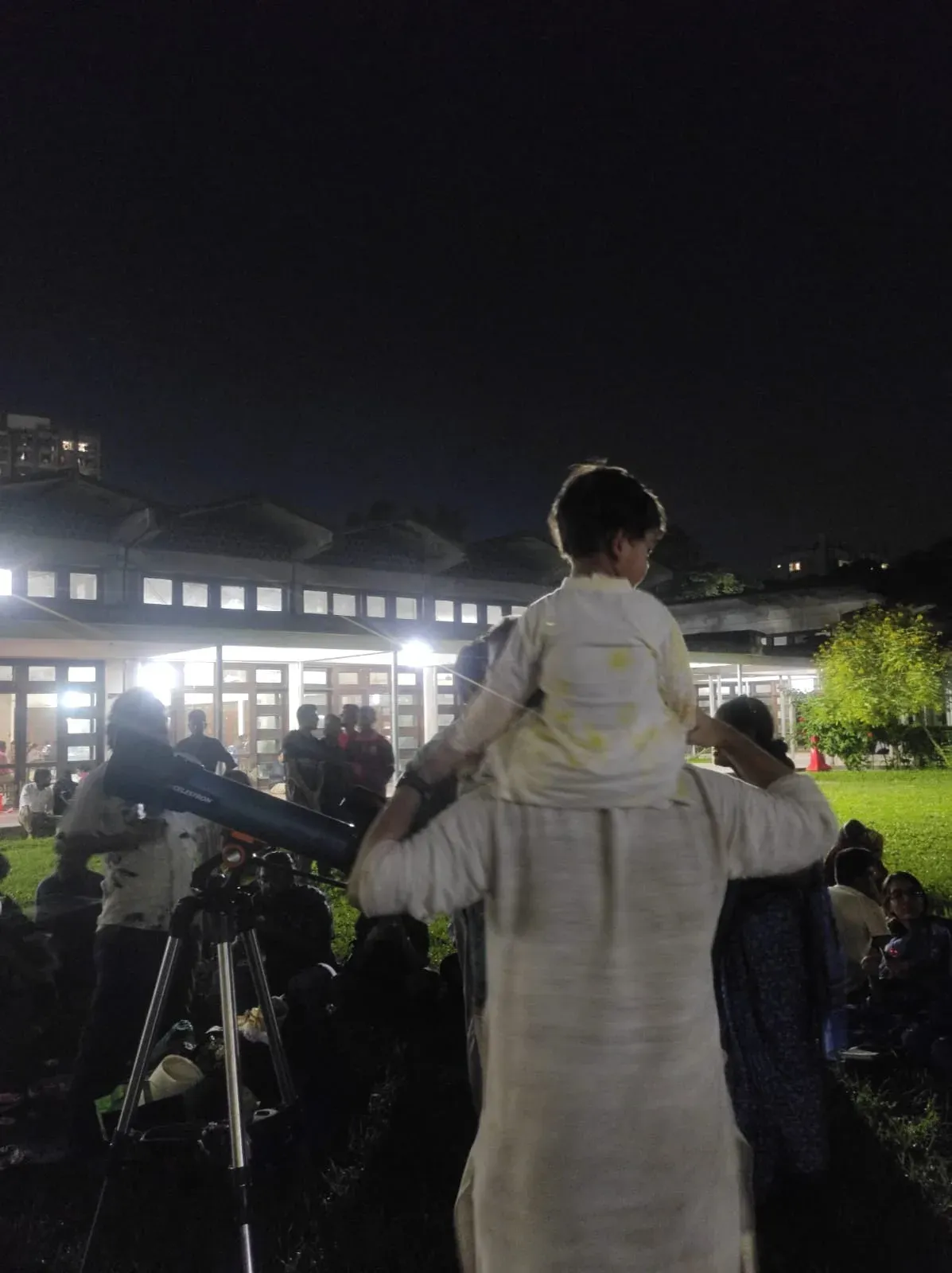 A child sitting on an adult's shoulders beside a Celestron telescope, looking out over the gathering at night.