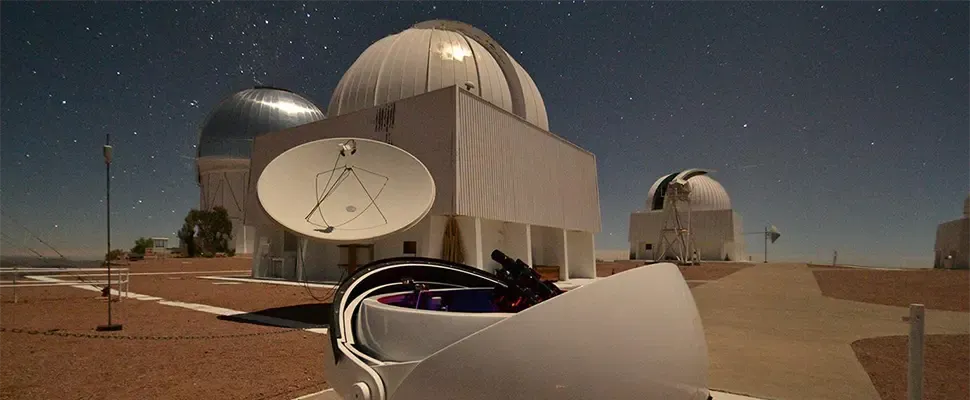 Telescope domes and a radio dish at a cosmological-survey observatory under a starry twilight sky.