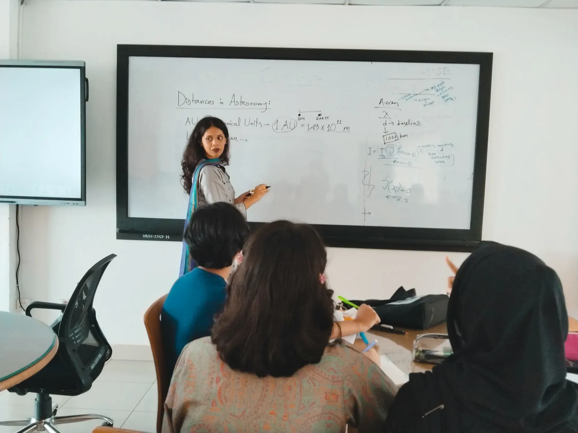 An instructor at the whiteboard beside the heading "Distances in Astronomy", with the astronomical unit written out as 1 AU = 1.49 × 10^11 m.