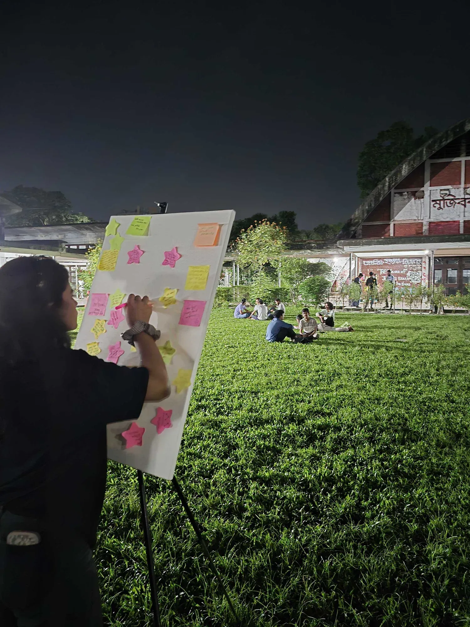 A volunteer writing on a canvas board covered in brightly coloured star-shaped sticky notes, with participants seated on the lawn behind.