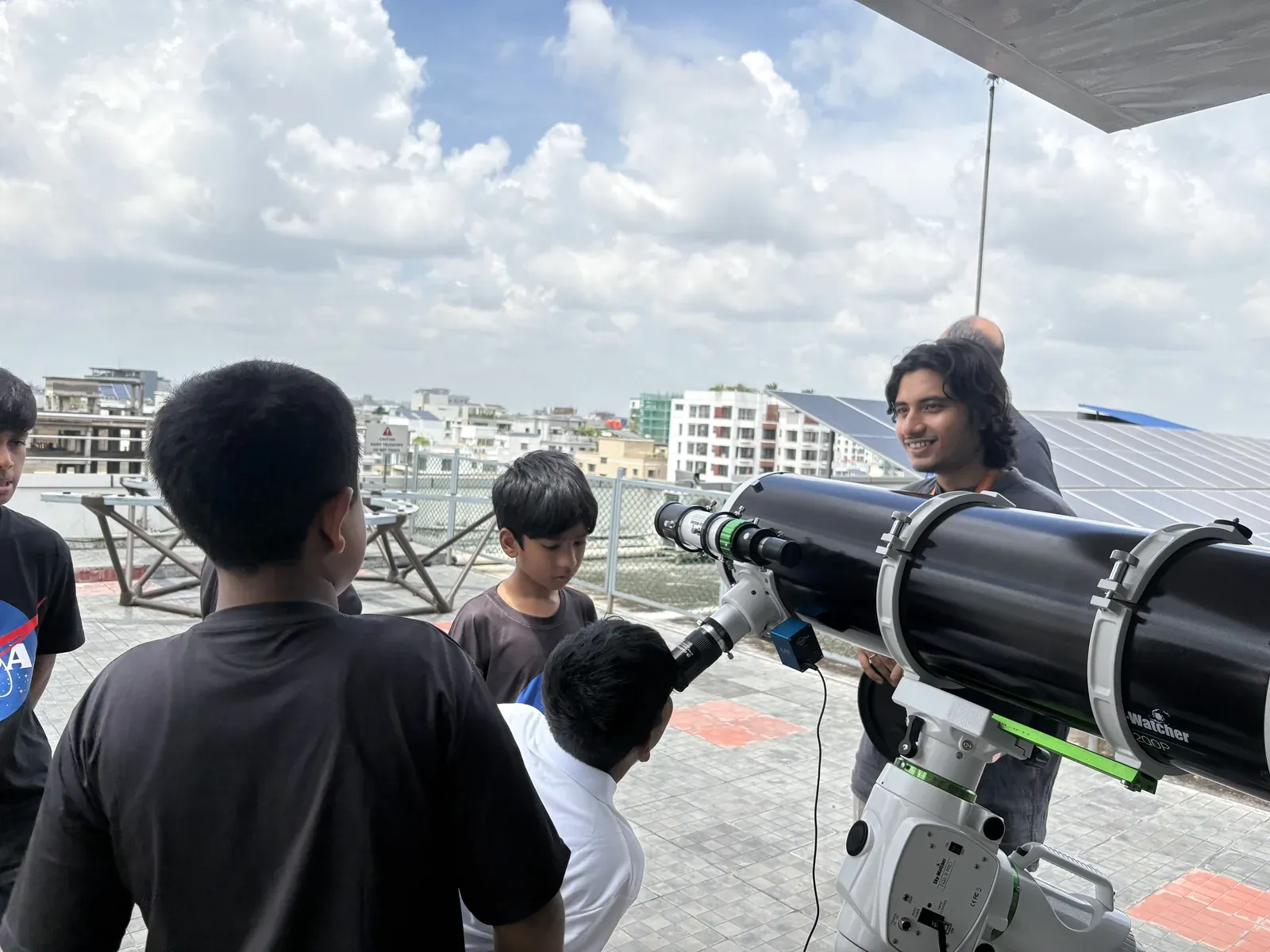 Md Shahadat Hossain Shahal stands behind the 8-inch Skywatcher telescope, smiling, as students look on.